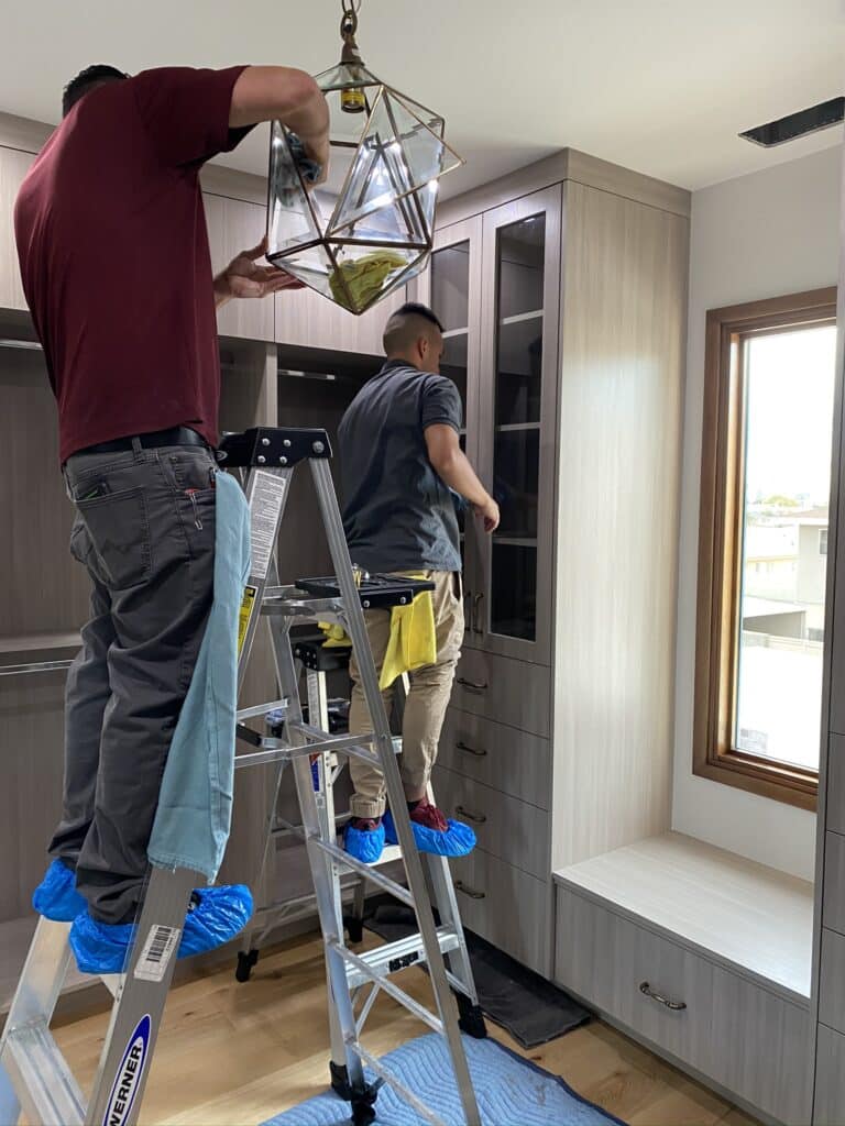 Two Nancie Brown crew members cleaning a chandelier and cabinetry during a post-construction cleanup in a modern home.